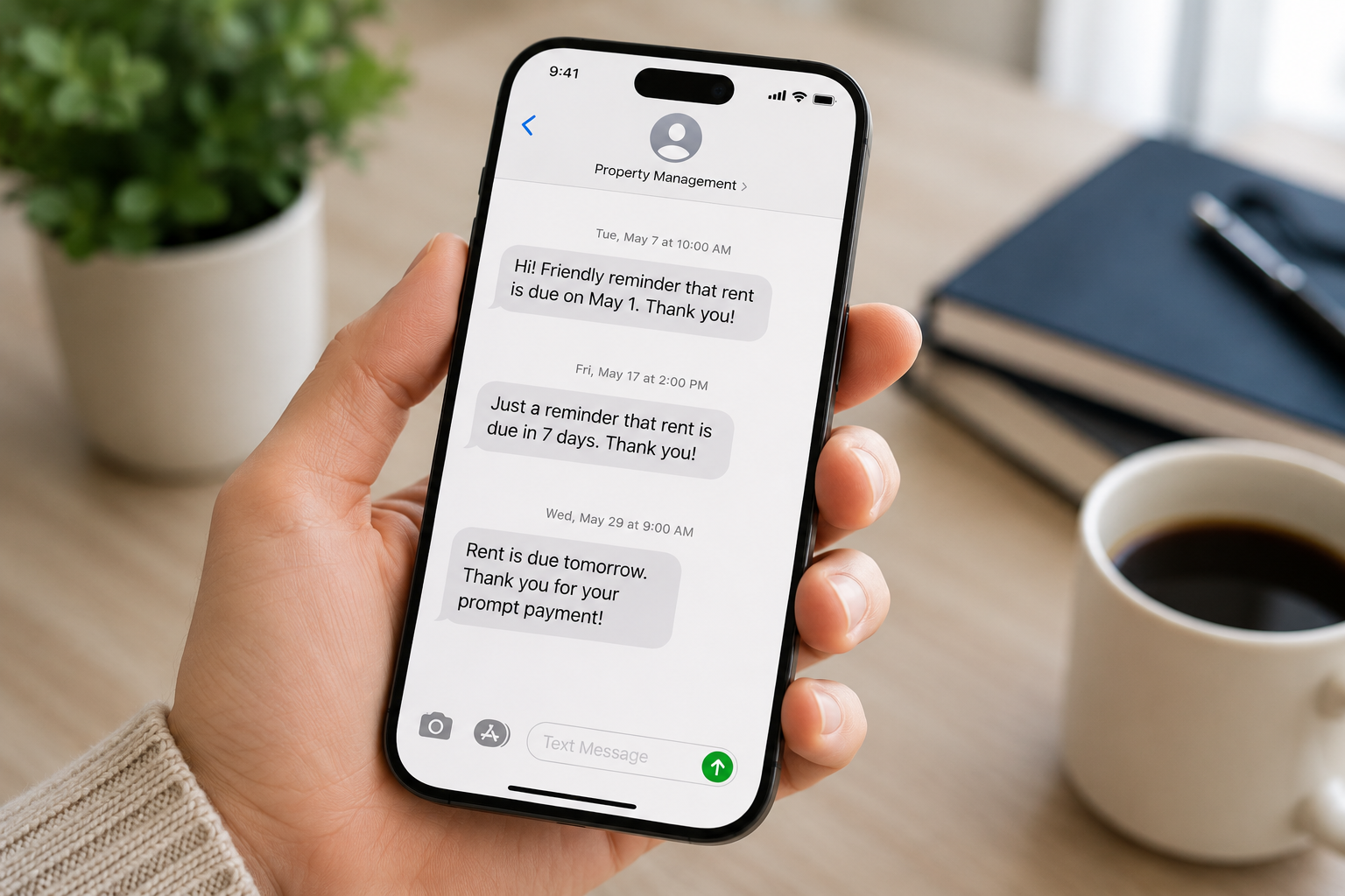A hand holds a smartphone displaying rent reminders from Property Management. A notebook and a coffee cup are on the wooden table in the background
